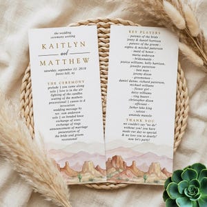 May include: Two vertical wedding program cards featuring gold text and a watercolor desert landscape illustration at the bottom. The text details the ceremony order, wedding party members, and a thank you note for Kaitlyn and Matthew's wedding.