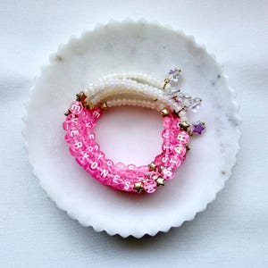 May include: A stack of three beaded bracelets featuring translucent pink and iridescent white beads. The pink beads spell out the phrase EVERYONES A STAR in white letters. Small gold-toned star charms and clear star-shaped pendants are attached.