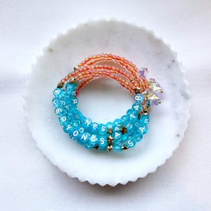 May include: A stack of beaded bracelets featuring translucent blue beads with white letters spelling EVERYONES A STAR, interspersed with small gold-tone star charms. The bracelets also include sections of iridescent orange beads and clear star-shaped accents.