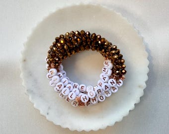 May include: A stack of three bracelets featuring a combination of metallic bronze faceted beads and white circular beads with rose gold lettering. The white beads spell out NIALL HORAN and include small rose gold star-shaped spacer beads.