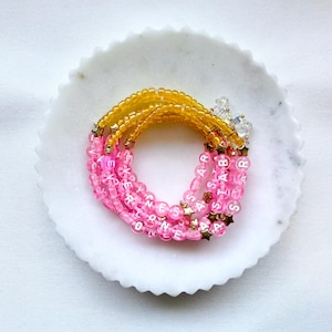 May include: A circular beaded bracelet featuring translucent pink beads with white letters spelling out EVERYONE IS A STAR, interspersed with small gold-toned star charms. The bracelet also includes a section of translucent yellow beads.