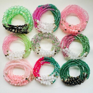 May include: Nine beaded bracelets in shades of green, pink, and white. Some bracelets have star-shaped beads and lettered beads spelling out names. The bracelets are arranged in a circular pattern, highlighting their design and colour variations.