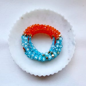 May include: A coiled bracelet made of faceted orange beads and translucent light blue beads. The blue beads feature white lettered cubes spelling out EVERYONES A STAR, interspersed with small gold-toned star-shaped metal spacers.