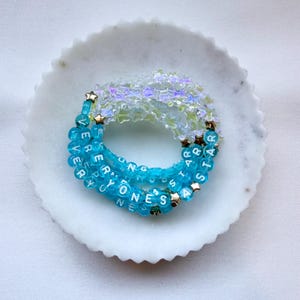 May include: A circular beaded bracelet featuring translucent star-shaped beads and bright blue spherical beads with white lettering that spells out EVERYONES A STAR. Small metallic gold star accents are interspersed throughout the design.
