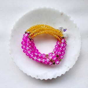 May include: A multi-strand beaded bracelet featuring vibrant yellow and hot pink translucent beads. The pink beads are interspersed with white letter beads spelling out EVERYONES A STAR, accented by small gold-toned star-shaped spacers.