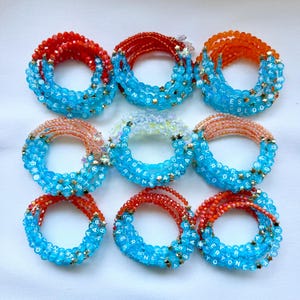 May include: Nine circular beaded bracelets arranged in a grid. Each bracelet features a combination of translucent light blue letter beads spelling EVERYONE IS A STAR, interspersed with small gold star charms and vibrant red or orange beads.