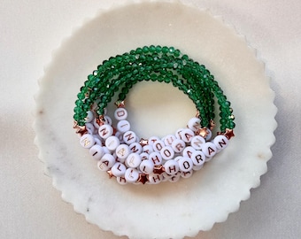 May include: A stack of three bracelets made of faceted emerald green beads and white round alphabet beads spelling NIALL HORAN. The bracelets feature small metallic copper-colored star-shaped spacers interspersed between the beads.