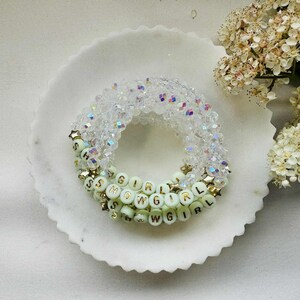 May include: Three beaded bracelets. The top bracelet is clear with iridescent beads. The middle bracelet is a light green with the words "SHOWGIRL" spelled out in white beads. The bottom bracelet is also light green with the words "SHOWGIRL" spelled out in white beads. Gold star accents.