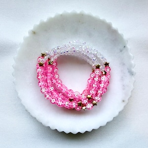 May include: A stack of beaded bracelets featuring translucent pink beads, clear iridescent beads, and small gold-toned star charms. The beads are imprinted with white letters spelling out the phrase EVERYONES A STAR, arranged in a circular pattern.