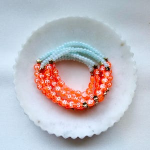 May include: A stack of three beaded bracelets featuring light blue beads on one side and translucent orange beads on the other. The orange beads are printed with white letters spelling out EVERYONES A STAR, accented with small gold-toned metal stars.