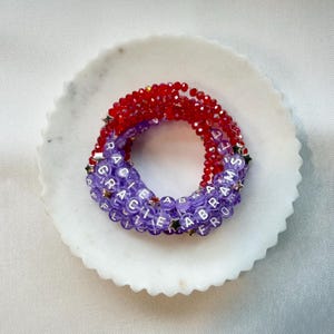 May include: A stack of beaded bracelets featuring faceted red beads and translucent purple beads with white lettered cubes spelling GRACIE ABRAMS. The bracelets are accented with small silver-toned star-shaped beads throughout the circular design.