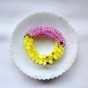 May include: A multi-strand beaded bracelet featuring a section of faceted pink beads and a section of bright yellow beads. The yellow beads are interspersed with small gold-toned star charms and white letter beads spelling out EVERYONES STAR.