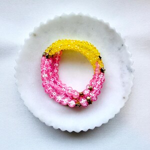 May include: A multi-strand beaded bracelet featuring bright yellow faceted beads and translucent pink beads with white lettered cubes spelling EVERYONES A STAR. Small gold-toned star-shaped spacers are interspersed throughout the design.