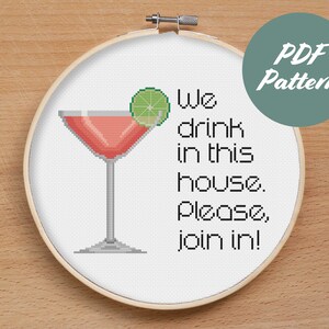 May include: A white embroidery hoop with a cross-stitch design of a pink cocktail with a lime wedge and the text "We drink in this house. Please, join in!"  The hoop is on a wooden surface.  PDF Pattern is written in green on a teal circle.