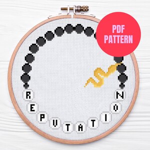 May include: A cross-stitch pattern in a wooden embroidery hoop. The design features the word "REPUTATION" in white letters, a black beaded border, and a yellow snake. A red circle with "PDF PATTERN" is in the upper right.