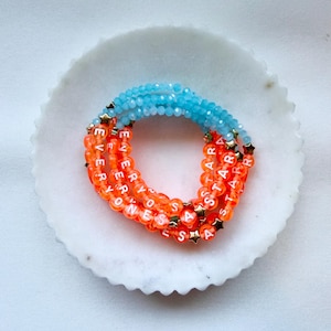 May include: A stack of beaded bracelets featuring bright orange round beads with white lettered text that reads EVERYONES A STAR, interspersed with small gold-toned star charms and a top section of translucent light blue faceted beads.