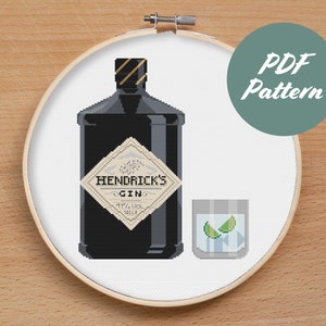 Könnte beinhalten: Ein Kreuzstichmuster für eine schwarze Flasche Hendrick's Gin mit Etikett und ein Glas Gin mit Limettenspalten. Das Muster befindet sich in einem weißen Stickrahmen auf einer Holzoberfläche. Der Text auf dem Etikett lautet "Hendrick's Gin 41,4% vol. 70cl."