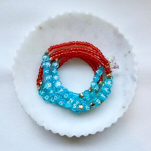 May include: A coiled bracelet made of small red beads and larger translucent light blue beads. The blue beads feature white letters spelling out EVERYONE IS A STAR, interspersed with small gold-toned star-shaped beads and a clear iridescent charm.