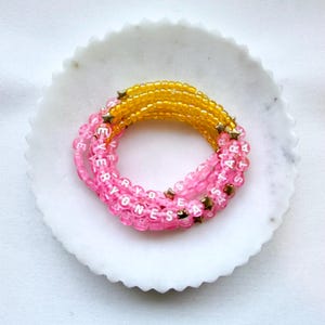 May include: A coiled bracelet made of translucent yellow and pink beads. The pink beads feature white letters spelling out EVERYONES A STAR, interspersed with small gold-coloured star-shaped beads throughout the circular design.