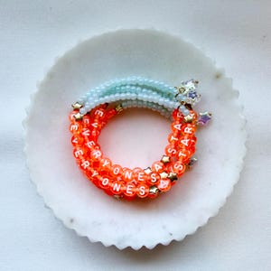 May include: A stack of beaded bracelets featuring light blue and bright orange translucent beads. The orange beads are printed with white letters spelling out EVERYONES A STAR, interspersed with small gold-toned star-shaped metal charms.