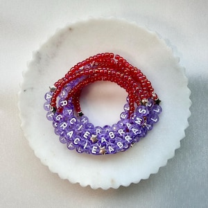 May include: A stack of beaded bracelets featuring translucent red and purple beads. The purple beads are interspersed with white letter beads spelling out GRACIE ABRAMS and small silver star-shaped beads, creating a layered accessory design.