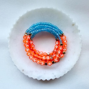 May include: A multi-strand beaded bracelet featuring translucent blue beads on the top half and vibrant orange beads on the bottom half. The orange section includes white letter beads spelling out EVERYONES A STAR, interspersed with small gold star charms.