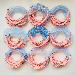 May include: Nine beaded bracelets in pink and blue. Each bracelet has letter beads spelling out names, with star-shaped accents. The bracelets are arranged in a circular pattern.