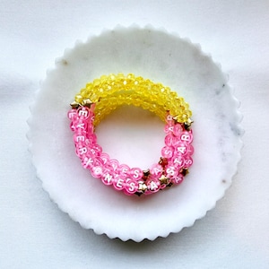 May include: A multi-strand bracelet featuring faceted yellow beads and translucent pink beads with white lettered cubes spelling EVERYONES A STAR, accented with small gold-toned star-shaped spacers throughout the circular design.