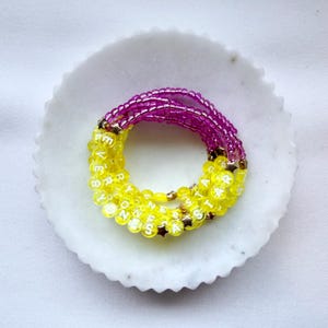 May include: A multi-strand bracelet featuring translucent yellow beads with white letters spelling EVERYONES A STAR, interspersed with small gold-toned star charms, and a section of vibrant purple beads, all arranged in a circular formation.