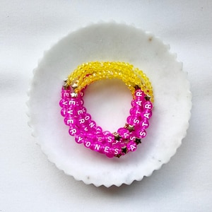 May include: A stack of beaded bracelets featuring bright yellow faceted beads and vibrant pink round beads. The pink beads are imprinted with white letters spelling out EVERYONES A STAR, interspersed with small metallic gold star-shaped beads.