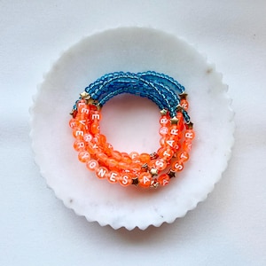 May include: A stack of beaded bracelets featuring translucent orange beads with white lettered beads spelling EVERYONES A STAR and translucent blue beads. Small gold-toned star-shaped spacers are interspersed throughout the bracelet design.