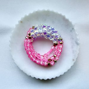May include: A circular beaded bracelet featuring translucent iridescent star-shaped beads and round pink beads with white lettering that spells out EVERYONES A STAR, accented with small gold-toned star beads throughout the design.