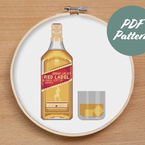 Puede incluir: Patrón de punto de cruz para una botella de whisky escocés Johnnie Walker Red Label y un vaso de whisky con cubitos de hielo. El patrón está en un aro de bordado blanco sobre una superficie de madera.