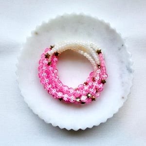 May include: A stack of three beaded bracelets featuring translucent pink beads, white beads with the black text EVERYONE IS A STAR, and small metallic gold star-shaped spacers. The bracelets are arranged in a circular pattern on a white surface.
