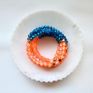 May include: A stack of three beaded bracelets featuring faceted blue beads, translucent orange beads with white lettered text reading EVERYONES A STAR, and small gold-toned star-shaped spacers arranged in a circular pattern on a white scalloped dish.