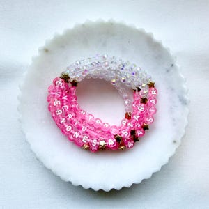 May include: A coiled bracelet made of translucent pink and clear faceted beads. The pink beads feature white letters spelling out EVERYONES A STAR, interspersed with small gold-toned star-shaped charms throughout the circular design.