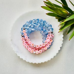 May include: Two beaded bracelets, one light blue and one pink. The pink bracelet spells "OLIVIA RODRIGO" in gold letter beads, with silver star beads. The bracelets are displayed on a white marble plate.