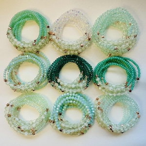 May include: Nine coiled beaded bracelets in shades of green and white. Each bracelet features small beads, star-shaped accents, and letters. The bracelets vary in colour from light green to dark green. The bracelets are arranged in a grid.