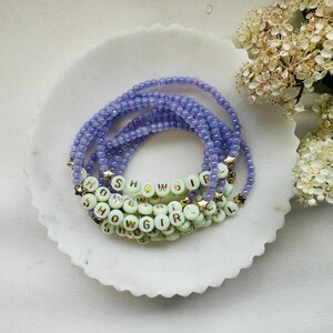 May include: Stack of lavender beaded bracelets with the word "SHOWGIRL" spelled out in white letter beads. Gold star-shaped beads accent the bracelets. The bracelets are arranged on a white, scalloped-edge dish.
