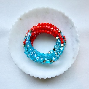 May include: A multi-strand beaded bracelet featuring vibrant red faceted beads and translucent light blue beads. The blue beads are interspersed with small gold-toned star charms and white letter beads that spell out the phrase EVERYONE IS A STAR.