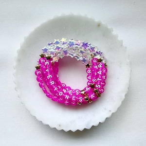 May include: A circular arrangement of beaded bracelets featuring vibrant hot pink spherical beads with white lettered text spelling EVERYONES A STAR, interspersed with small gold-toned star charms and translucent iridescent star-shaped beads.