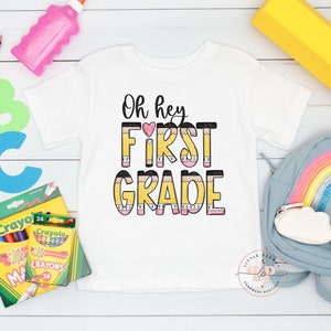 May include: A white t-shirt with the text "Oh hey FIRST GRADE" in black and pink letters with a yellow pencil design. The shirt is surrounded by school supplies, including crayons, a pencil, a glue stick, a backpack, and a large yellow pencil.