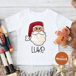 May include: White t-shirt with a cartoon image of Santa Claus and the name "Luke" printed on it. The shirt is decorated with a red and white candy cane design and a gingerbread man.