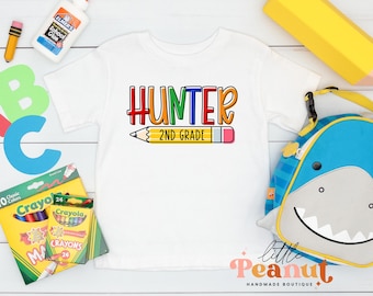 Può includere: Maglietta bianca con il testo "HUNTER 2ND GRADE" in lettere colorate e un'immagine di una matita arcobaleno. La maglietta è circondata da materiale scolastico, tra cui matite colorate, colla e una matita.