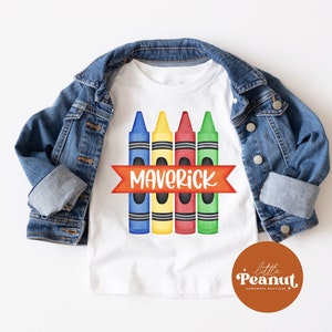May include: White t-shirt with a design of four colorful crayons and a red banner with the name "Maverick" on it. The t-shirt is folded and displayed on a white background with a denim jacket.