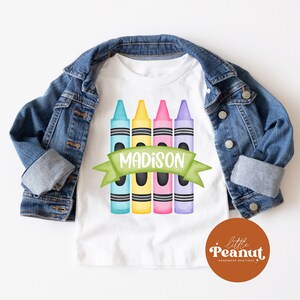 May include: White t-shirt with a design of five colorful crayons and a green banner with the name "MADISON" on it.