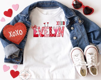 May include: White t-shirt with a red, pink, and green heart design that spells out the name "EVELYN" with the letters "XOXO" above the name. The shirt is laid out on a white surface with a denim jacket, red heart-shaped sunglasses, and red and pink hearts.
