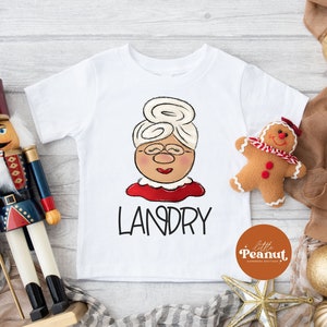 May include: White t-shirt featuring a cartoon illustration of Mrs. Claus with glasses, a red collar, and the name "LANDRY" in black. A gingerbread man and a nutcracker are next to the shirt. The shirt has a logo that says "Little Peanut Handmade Boutique."