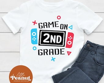 Può includere: Maglietta bianca con un design grafico di un controller per videogiochi con il testo "Game On 2nd Grade".