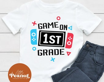 May include: White t-shirt with a blue and red video game controller graphic and the text "Game On 1st Grade".